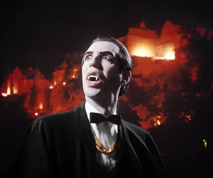 1970s MAN IN VAMPIRE MAKEUP AND COSTUME LOOKING UP MENACINGLY MOUTH OPEN SHOWING HIS BLOODTHIRSTY FANGS CASTLE BACKGROUND