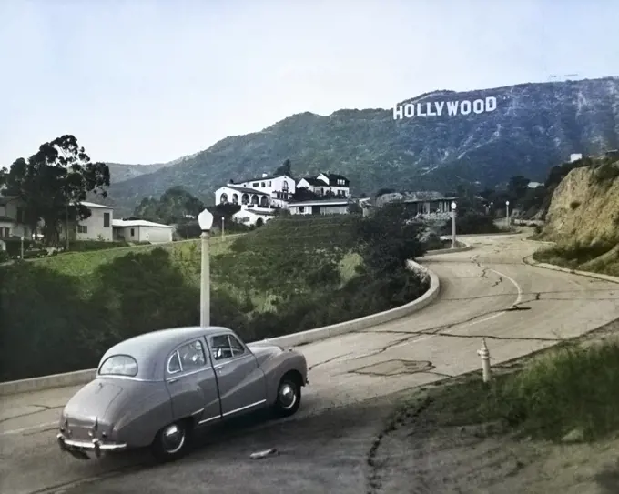 1950s AUSTIN CAR DRIVING UP ROAD IN THE HOLLYWOOD HILLS WITH HOLLYWOOD SIGN IN DISTANCE LOS ANGELES CALIFORNIA USA