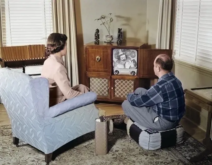 1950s MIDDLE AGE COUPLE HUSBAND AND WIFE SITTING WATCHING BLACK AND WHITE CONSOLE TELEVISION IN LIVING ROOM