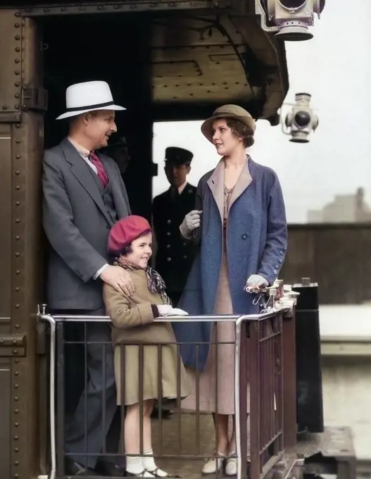 1930s FAMILY OF THREE STANDING ON BACK PLATFORM OF PASSENGER TRAIN PARLOR OBSERVATION CAR WITH CONDUCTOR TRAINMAN IN BACKGROUND