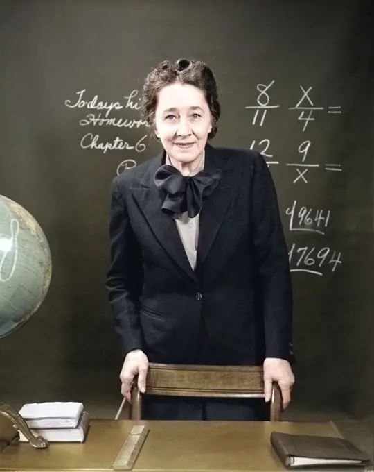 1940s SMILING SCHOOL TEACHER STANDING BEHIND HER DESK & IN FRONT OF THE BLACK BOARD WITH FRACTIONS