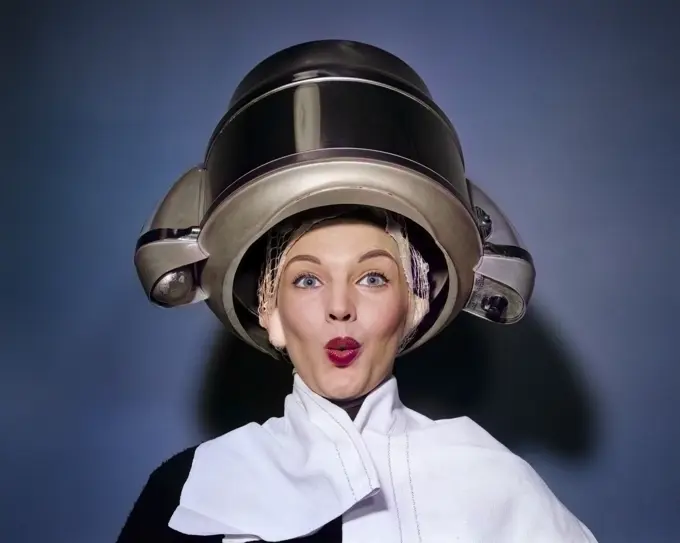 1950s SURPRISED WOMAN SITTING UNDER HAIR DRYER WITH TOWEL ON SHOULDERS AND HAIR NET LOOKING AT CAMERA