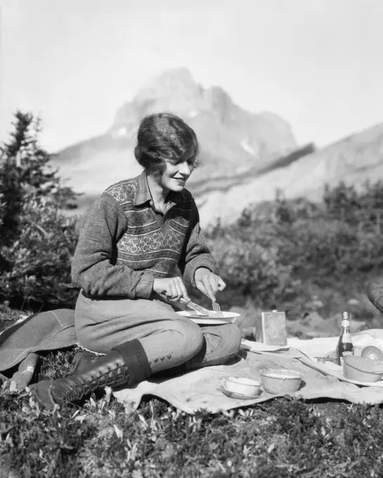 1920s WOMAN CAMPING EATING BREAKFAST OUTDOORS SITTING ON GRASS MOUNTAINS IN BACKGROUND BAKER LAKE ALBERTA CANADA