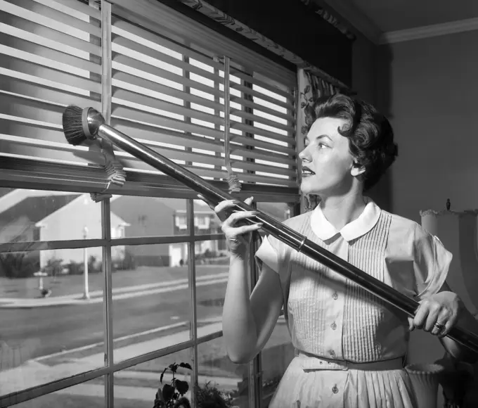 1950s WOMAN VACUUMING VENETIAN BLINDS ON WINDOW LOOKING OUT ONTO SUBURBAN STREET