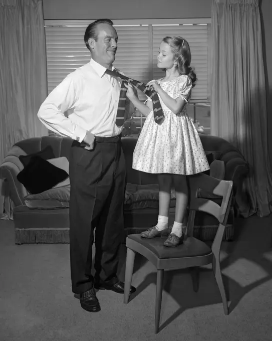 1950s CONFIDENT LITTLE GIRL STANDING ON CHAIR TYING NECKTIE FOR HER SMILING FATHER