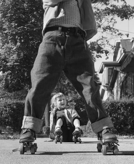 1950s GIRL FALLEN SITTING ON SIDEWALK WEARING METAL ROLLER SKATES SHOT THROUGH LEGS OF BOY ROLLED CUFF BLUE JEANS