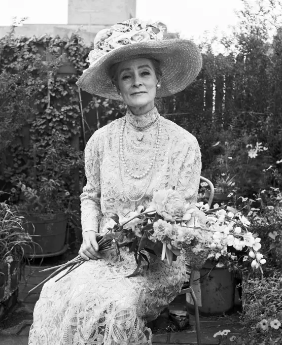 1970s SENIOR WOMAN SITTING IN GARDEN HOLDING BOUQUET FLOWERS WEARING ANTIQUE LACE DRESS BIG HAT SMILING LOOKING AT CAMERA