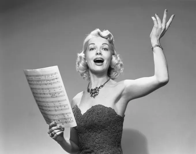 1950s BLOND WOMAN SINGING HOLDING SHEET MUSIC WEARING STRAPLESS DRESS NECKLACE Photographer: H. ARMSTRONG ROBERTS