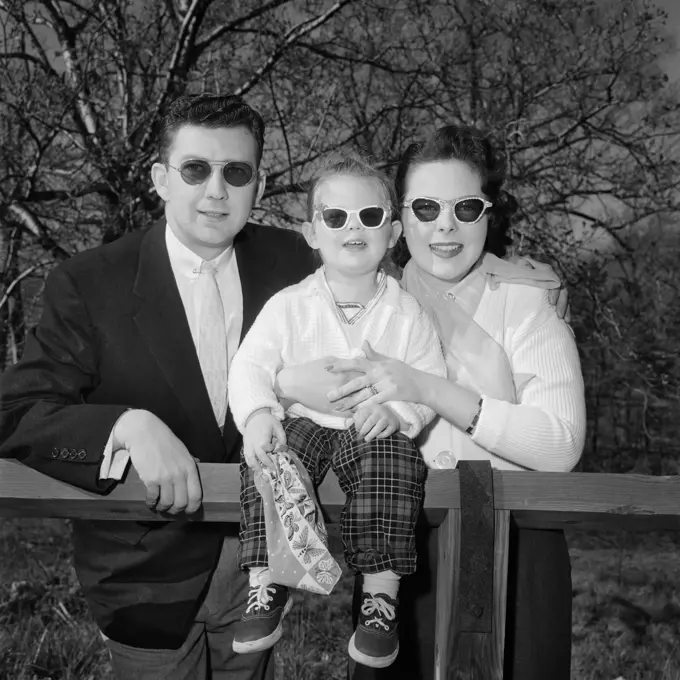 1950s FAMILY FATHER MOTHER DAUGHTER AT FENCE WEARING DARK SUNGLASSES LOOKING AT CAMERA