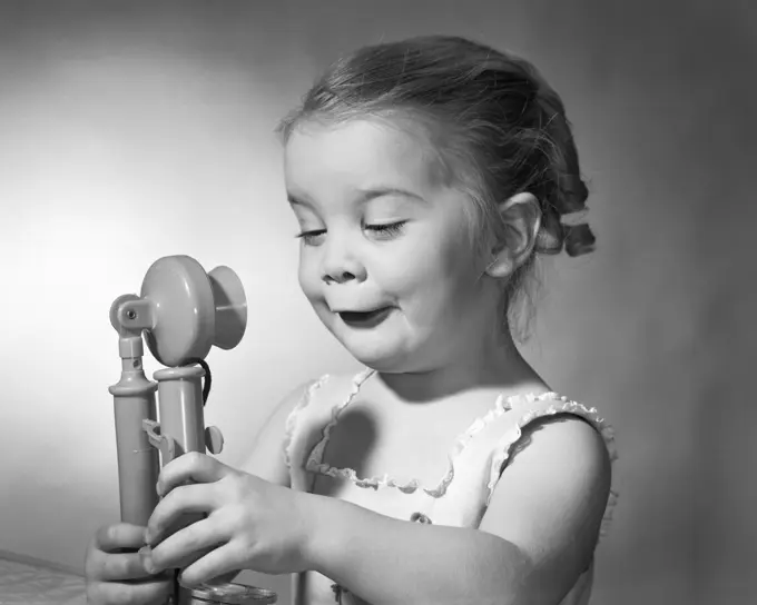 1960s LITTLE GIRL WITH FUNNY FACIAL EXPRESSION HOLDING A TOY CANDLE STICK TELEPHONE IN BOTH HANDS