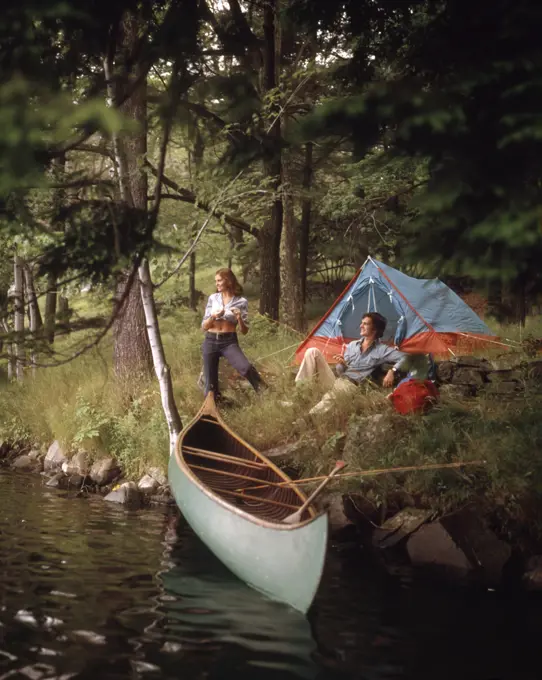 1970s COUPLE AT A FOREST CAMP CAMPSITE WITH TENT AND CANOE IN STREAM 