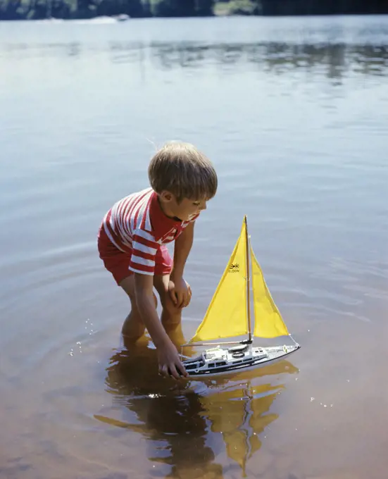 1970s BOY WADING IN POND WATER PLAYING WITH TOY BOAT WITH YELLOW SAILS 