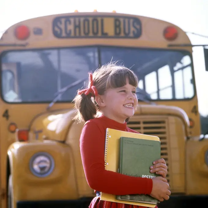 1970s SMILING ELEMENTARY SCHOOL GIRL STANDING FRONT OF BUS CARRYING BOOKS SMILING 