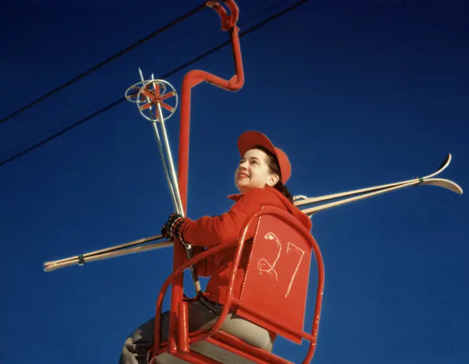 1950s BRUNETTE WOMAN RED SKI HAT JACKET SITTING IN RED SKI LIFT CHAIR HOLDING VINTAGE SKIS SKI POLES
