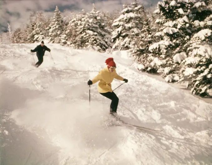 1960s 1970s TWO PEOPLE MAN WOMAN SKIING DOWN SNOWY SLOPE PINE TREES WOMAN WEARING YELLOW JACKET AND RED CAP