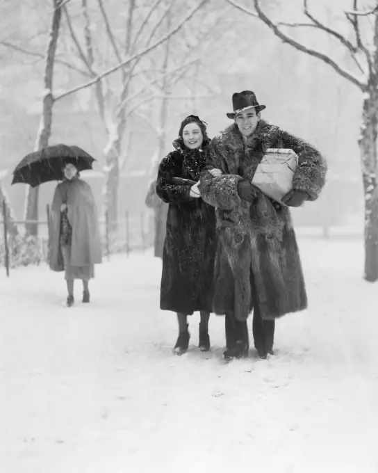 1930s SMILING CHRISTMAS SHOPPING COUPLE WEARING FUR COATS WALKING THROUGH SNOWY CITY PARK CARRYING PACKAGES LOOKING AT CAMERA