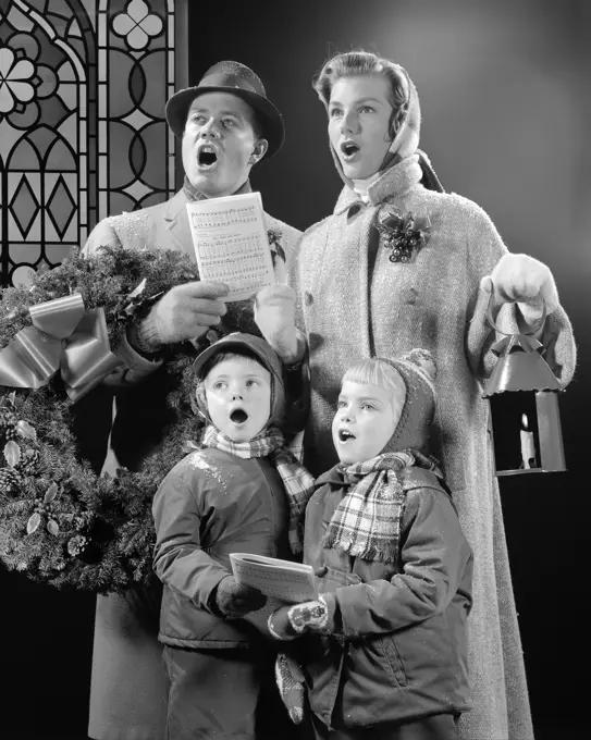 1950s FAMILY OF FOUR SON DAUGHTER SINGING CHRISTMAS CAROLS DAD HOLDING WREATH MOM HOLDING CANDLE LANTERN