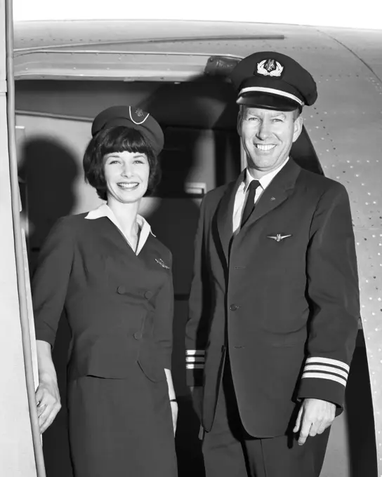 1960s MAN CO-PILOT AND WOMAN FLIGHT ATTENDANT STANDING SMILING IN DOOR OF AIRPLANE LOOKING AT CAMERA 