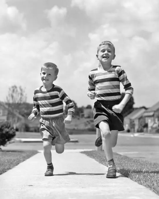 1950s TWO SMILING BOYS BROTHERS WEARING STRIPED SHIRTS SHORTS RUNNING ON SIDEWALK LOOKING AT CAMERA SUBURBAN NEIGHBORHOOD