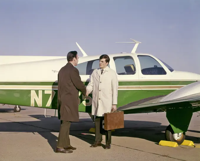 1960s TWO BUSINESS MEN SHAKING HANDS STANDING BESIDE SMALL PRIVATE AIRPLANE