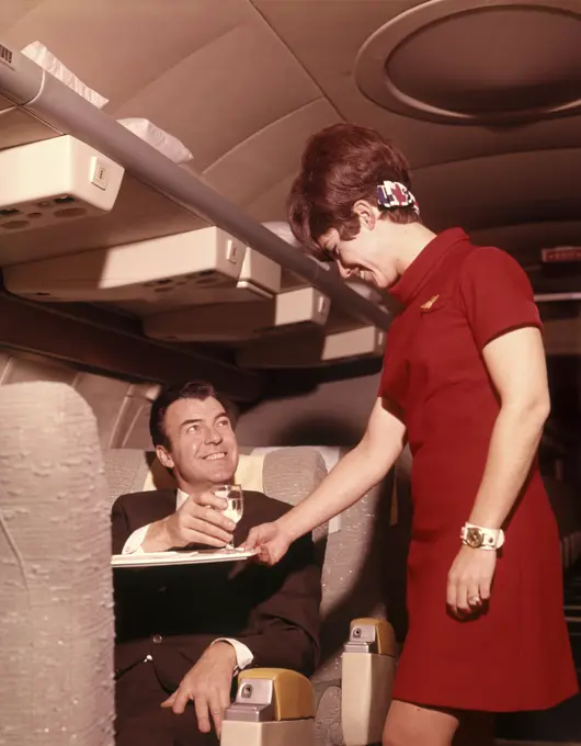 1960s 1970s AIRPLANE PLANE STEWARDESS SERVING BUSINESS MAN DRINK WINE WOMAN COMMERCIAL AIR TRAVEL