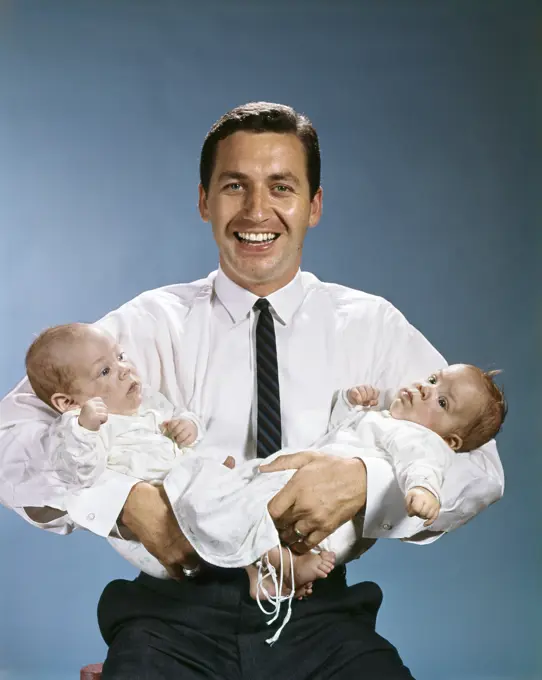 1960s SMILING FATHER LOOKING AT CAMERA HOLDING TWO TWIN BABIES IN ARMS