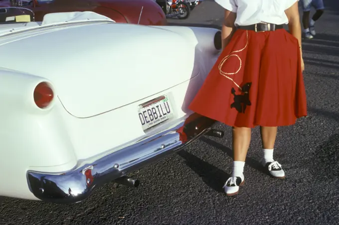 WOMAN IN RED POODLE SKIRT AND SADDLE SHOES NEXT TO WHITE 1950s CAR