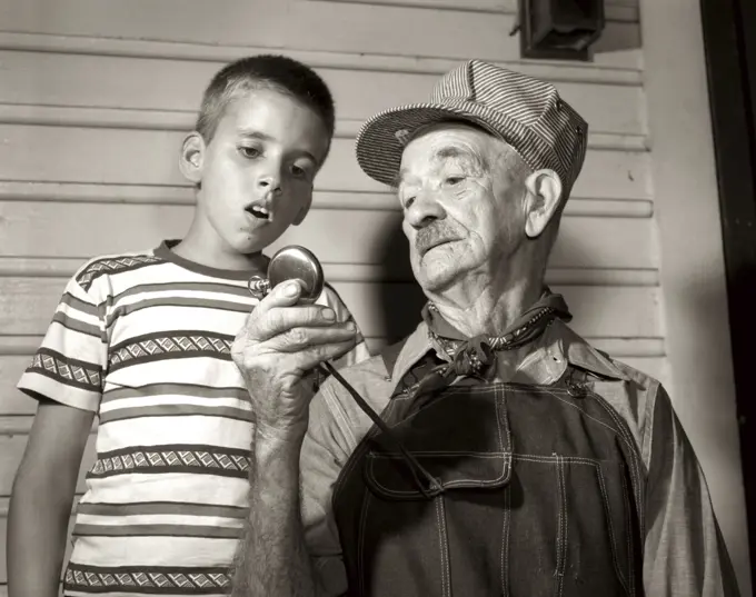 1950s ELDERLY MAN GRANDFATHER TRAIN ENGINEER HOLDING UP POCKET WATCH FOR BOY GRANDSON TO SEE