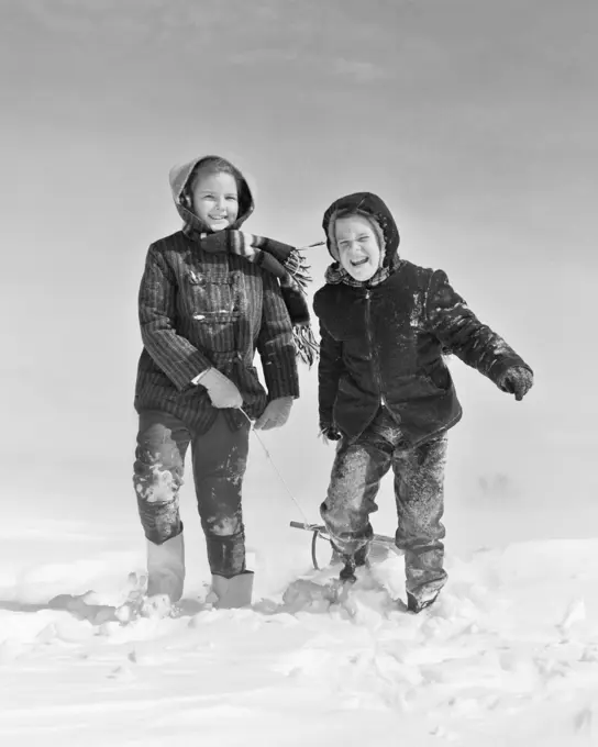 1950s 1960s SMILING BOY GIRL PULLING SLED IN WINTER SNOW LOOKING AT CAMERA