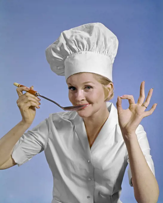 1960S WOMAN CHEF WEARING TOQUE LOOKING AT CAMERA TASTING FOOD MAKING OKAY SIGN SMILING