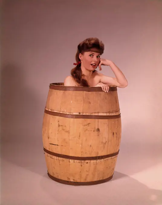 1960s YOUNG NUDE BRUNETTE WOMAN SITTING CROUCHING IN LARGE WOODEN BARREL LOOKING AT CAMERA