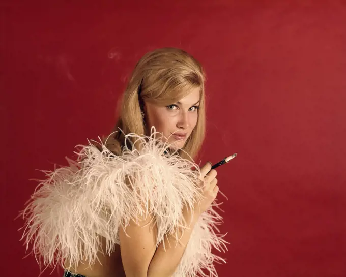 1960s SEXY BLONDE WOMAN LOOKING AT CAMERA SMOKING CIGARETTE IN LONG HOLDER WEARING WHITE FEATHER BOA AGAINST RED BACKDROP
