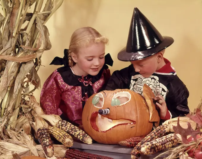 1960s TWO KIDS BLOND BROTHER AND SISTER WEARING HALLOWEEN COSTUMES LOOKING INTO CARVED JACK-O-LANTERN PUMPKIN