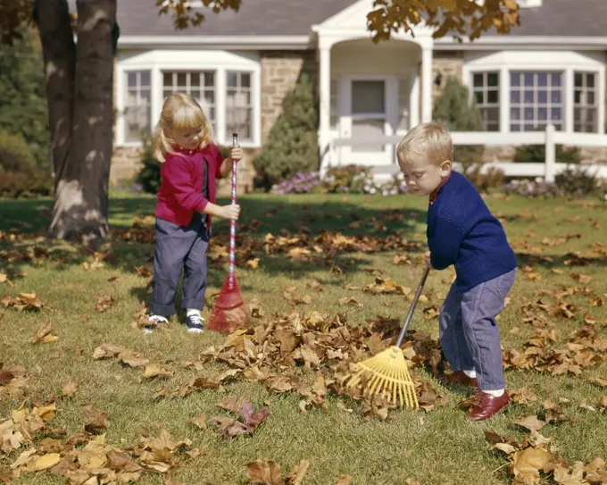 1960s BOY GIRL RAKING AUTUMN LEAVES IN YARD SUBURBAN HOUSE IN BACKGROUND 