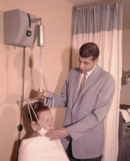 1950s 1960s MAN TECHNICIAN ADJUSTING TENSION ON WOMAN PATIENT CHIN BRACE NECK TRACTION MEDICAL EQUIPMENT