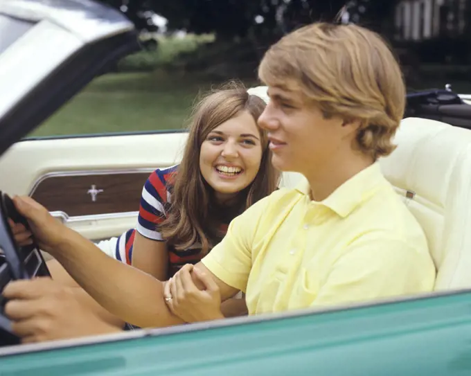 1960s 1970s SMILING TEENAGE COUPLE GIRL FLIRTING LOOKING AT HOLDING ARM OF BOY DRIVING CONVERTIBLE CAR 