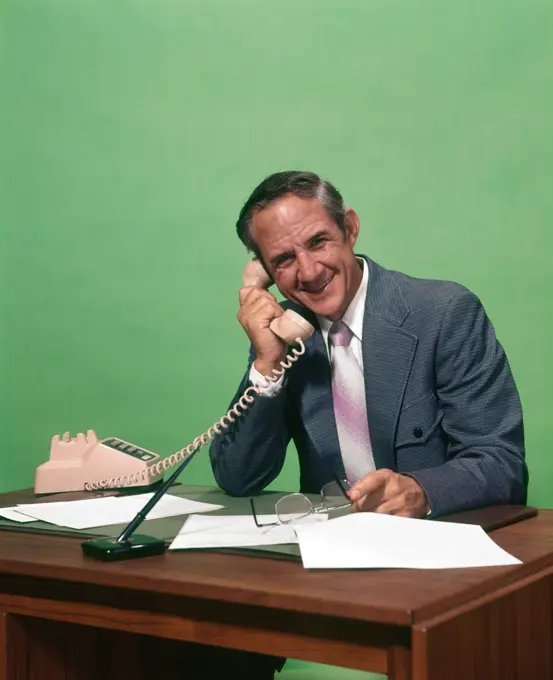 1970s SMILING MATURE MAN WEARING SUIT HOLDING GLASSES SITTING AT DESK SPEAKING ON TELEPHONE LOOKING AT CAMERA