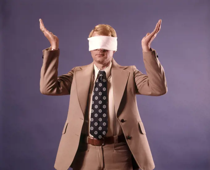 1970s EXECUTIVE MAN WEARING BUSINESS SUIT WEARING BLINDFOLD HOLDING BOTH HANDS UP IN HOPELESS GESTURE