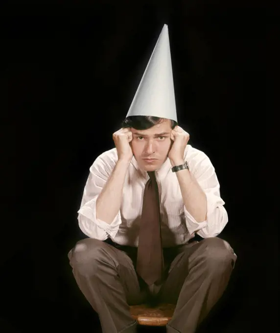 1960s UNHAPPY YOUNG MAN SITTING ON STOOL WEARING DUNCE CAP LOOKING AT CAMERA 