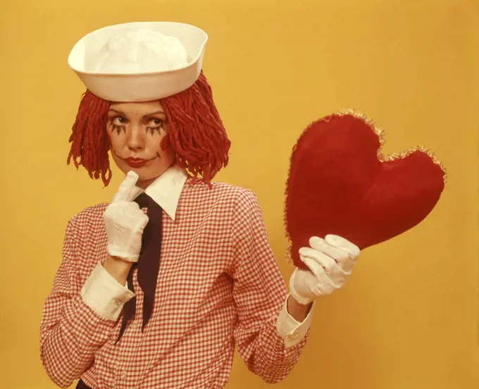 1970s YOUNG WOMAN RAGGEDY ANN WIG COSTUME SAILOR HAT YELLOW BACKGROUND HOLDING UP HEART
