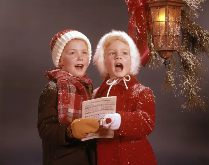 1960s TWO YOUNG CHILDREN BOY AND GIRL WINTER COATS SINGING CAROLS UNDER CHRISTMAS DECORATED CANDLE LANTERN