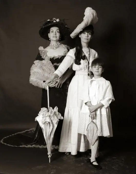 1960s THREE GENERATIONS WOMEN GRANDMOTHER MOTHER DAUGHTER POSED STANDING LOOKING AT CAMERA WEARING 1890s FASHION DRESSES