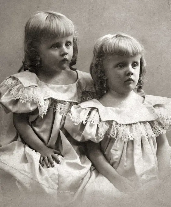 1890s PORTRAIT TURN OF THE 20TH CENTURY TWIN GIRLS SISTERS WEARING IDENTICAL DRESSES AND SHOWING MOROSE SAD FACIAL EXPRESSIONS
