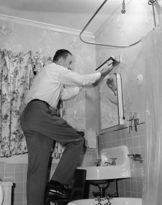 1950s MAN ON HOME REPAIR STEP STOOL IN BATHROOM REPLACING FLORESCENT LIGHTING FIXTURE ABOVE SINK