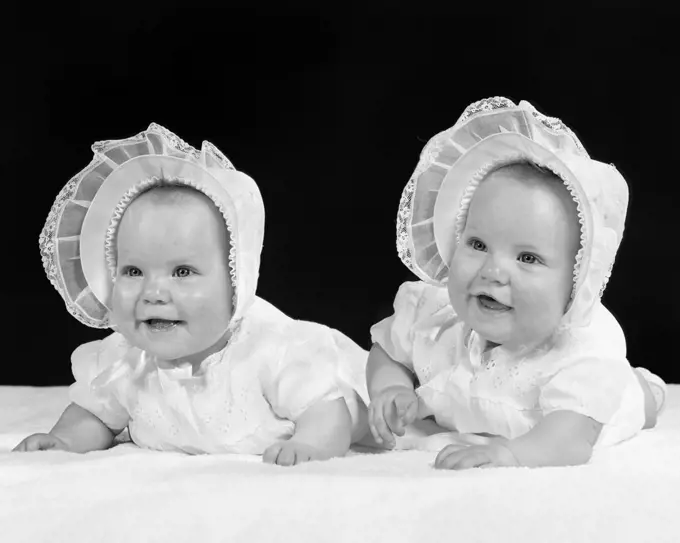 1950s TWIN BABY GIRLS WEARING BONNETS LYING ON THEIR STOMACHS SMILING
