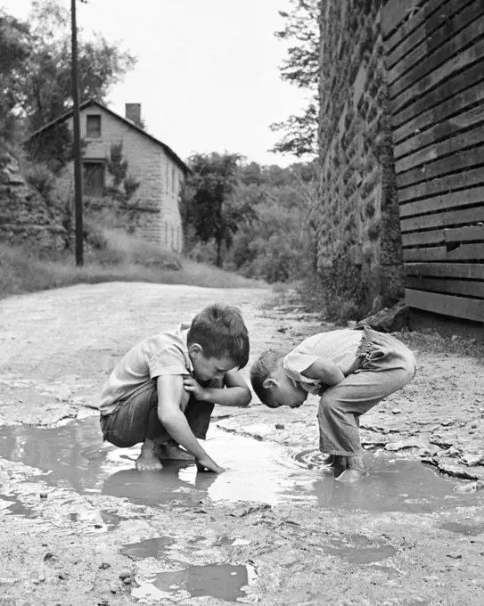 1950s 1960s TWO BOYS BENDING LOOKING AT PLAYING IN MUD PUDDLE