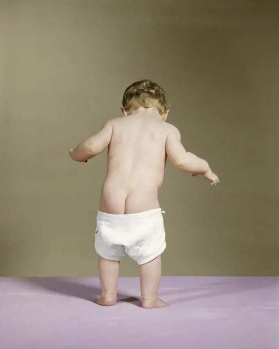 1960s REAR VIEW OF STANDING BABY WITH DIAPER FALLING DOWN SHOWING BARE BOTTOM