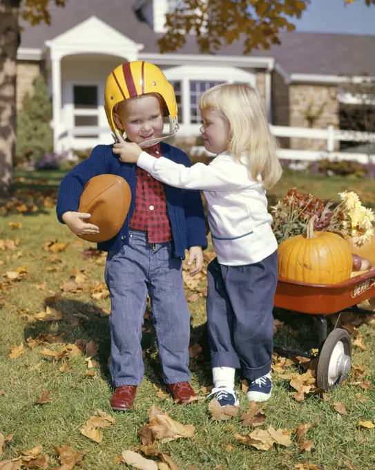 1960s BLONDE GIRL WEARING SADDLE OXFORDS HELPING BOY HOLDING FOOTBALL PUT ON HELMET AUTUMN YARD RED WAGON