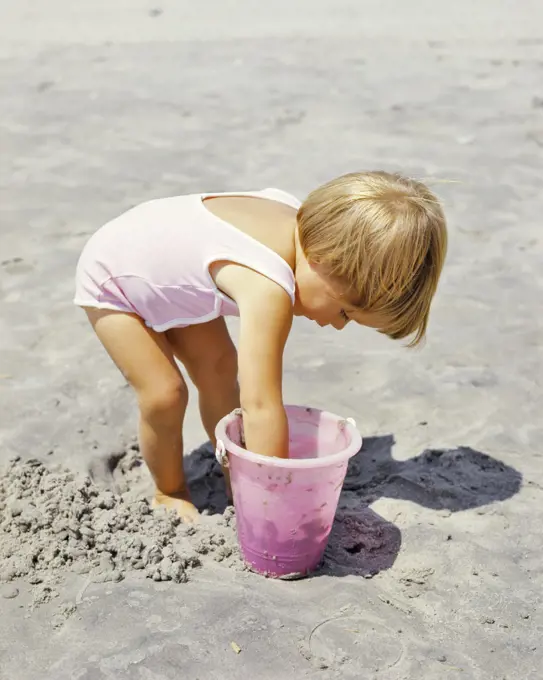 1970s LITTLE BLOND GIRL PINK BATHING SUIT PLAYING IN SAND WITH PINK PLASTIC PAIL ON BEACH