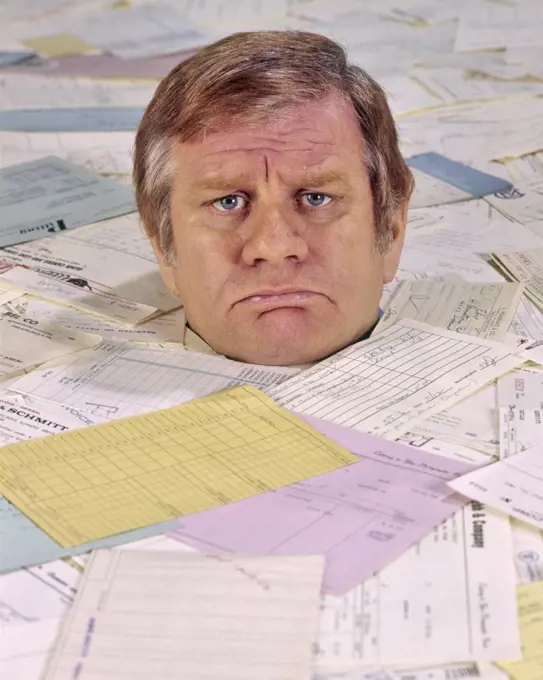 1970s 1980s ANGRY UNHAPPY FRUSTRATED MAN LOOKING AT CAMERA BURIED UP TO HIS NECK IN BILLS INVOICES PAPER WORK